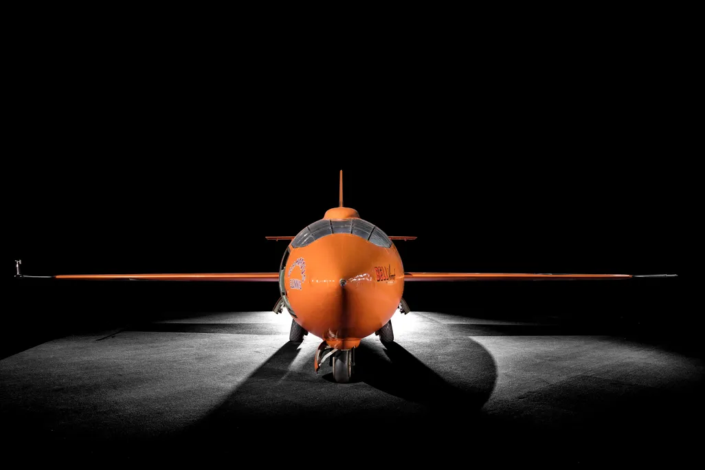 A bright orange plane, just big enough for one person to fit into, with cockpit facing the camera and each wing extended to either side, framed by dramatic lighting and a black background