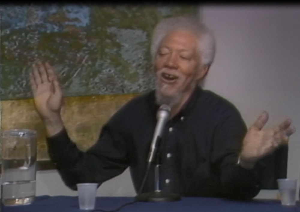 Benny Andrews sits at a table with a microphone, cups, and a pitcher of water with a yellow, green, and blue abstract painting hanging behind him. He is wearing a black button down shirt and raising his hands while talking.