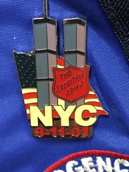 Pin featuring red salvation army seal and gray twin towers on top of American flag background