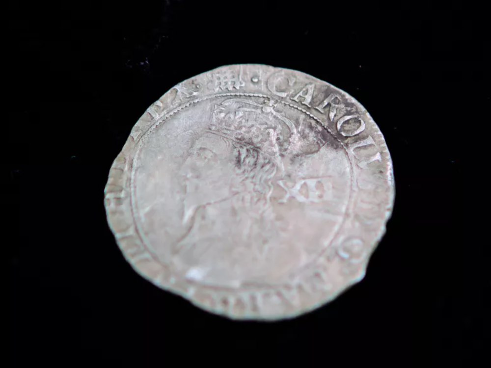 A silver coin with the face of a King and large capital letters going around in a circle; uneven edges