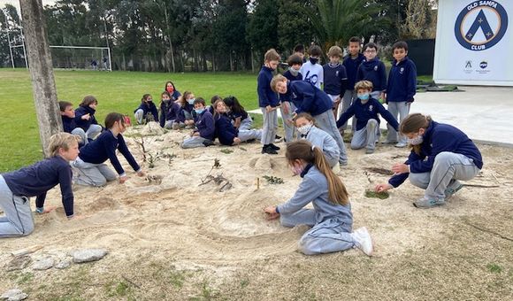Students cluster in groups and individually around a sandy area, adding and refining the community map they are building out of sand, leaves, and sticks.