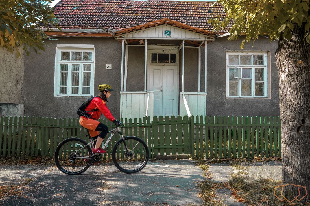 A young woman bikes in front of a traditional Armenian house.