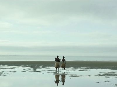 Two people in grass skirts stand on the shore, facing the ocean under an overcast sky.