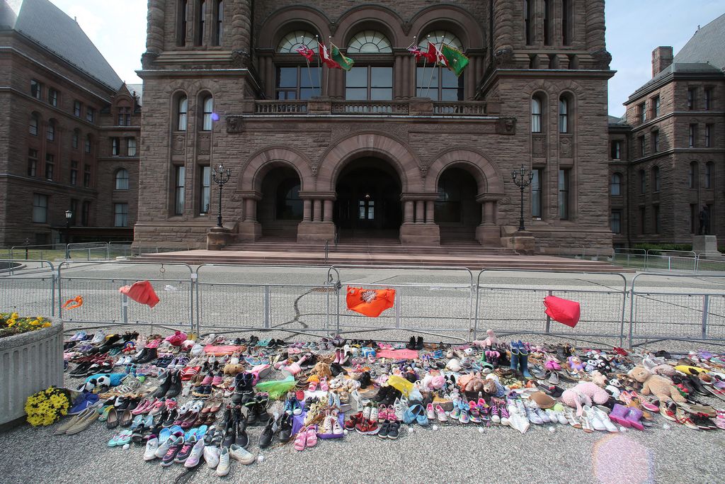 A memorial of children's shoes, all different colors, and flags are clustered on the concrete ground next to a fence, looking onto a big official building