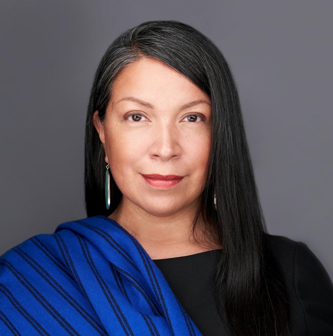 A headshot of Norby, who has black hair and wears a blue striped shawl over one shoulder, and faces the camera and smiles slightly