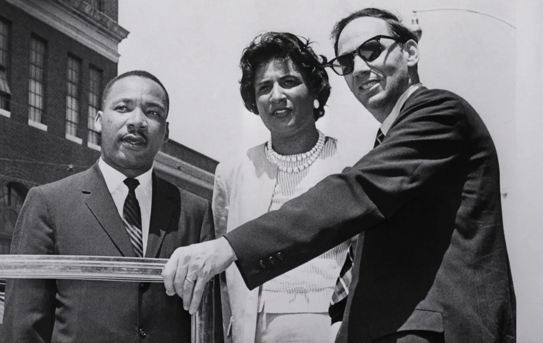A black and white image. Three people look out of frame as they enter a car, including Martin Luther King Jr, a Black man in suit and tie; Constance Baker Motley, a tall Black woman in pearls and sweater set; and William Kunstler, a white man in a suit