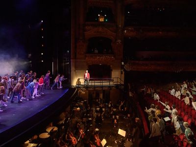 A performance in an opera hall, looking sideways across the orchestra pit. The performers are both on stage, lit in blue on the left, and in the first rows of red seats, on the right.
