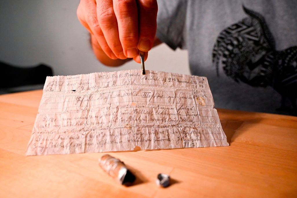 A person sits behind a table and delicately lifts the frail-looking slip of paper with tweezers; the sheet is thin and covered in spidery German cursive script; a small metal capsule that held the message lies on the table in front