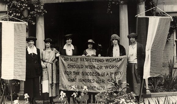Suffragists stand holding a banner quoting Susan B. Anthony: "No self respecting woman should wish or work for the success of a party that ignores her sex."