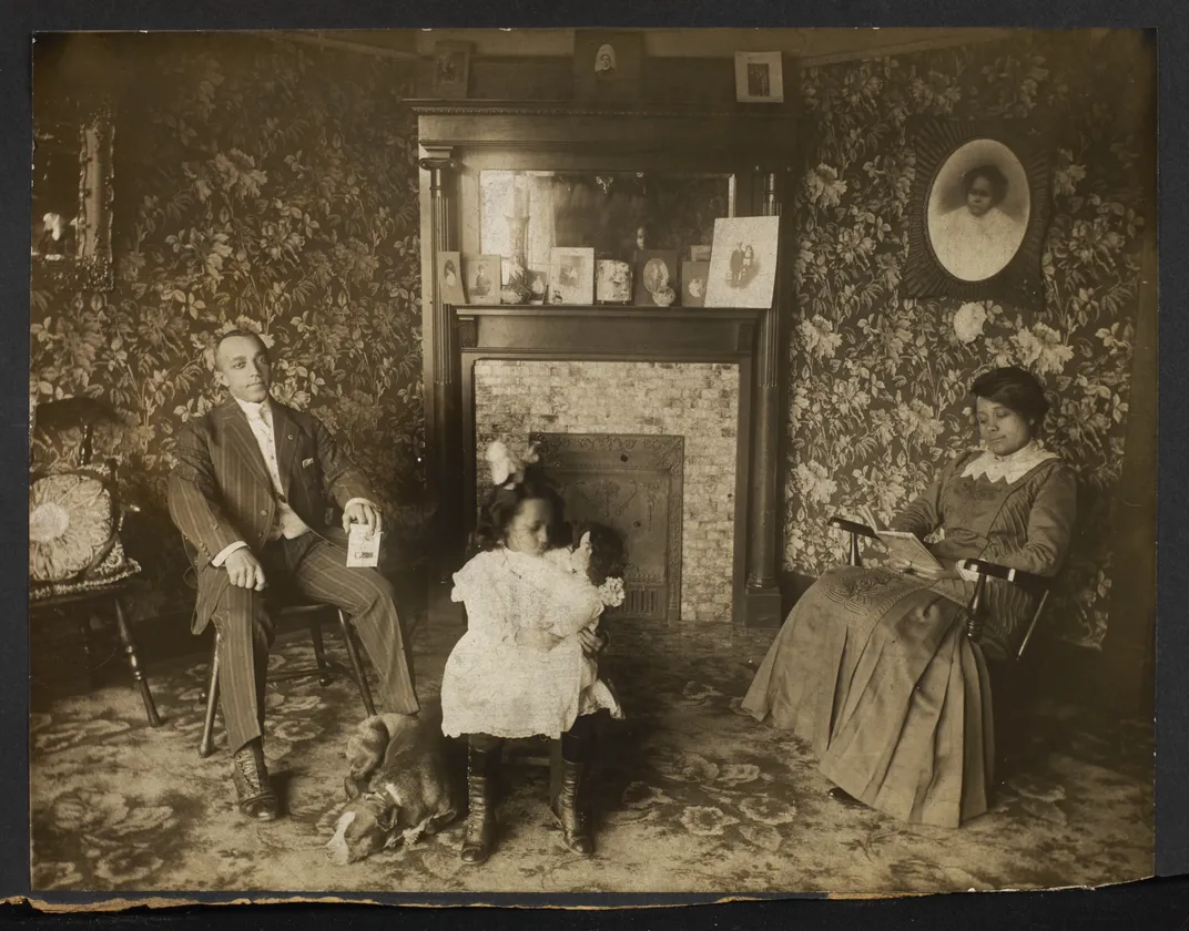 A sepia image of a middle-class family hearth, with a Black father, mother and child wearing fine clothes and posed around the fireplace; the child holds a White doll