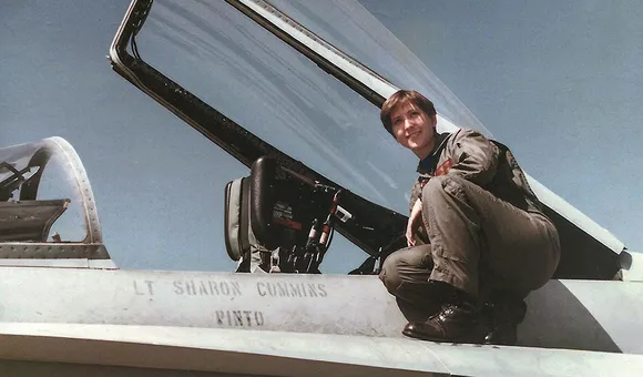A young woman wearing a U.S. Navy olive-green flightsuit and black combat boots perches next to the cockpit of her F/A-18C miltary jet. "Lt Sharon Cummins" is stenciled onto the side of the airplane.
