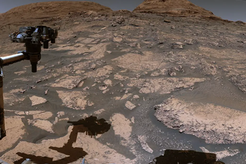 An image taken with the Curiosity Mars Rover. The image showed the Mars's dusty surface and a few hills in the background. The rover's arm is also seen in the photo. 