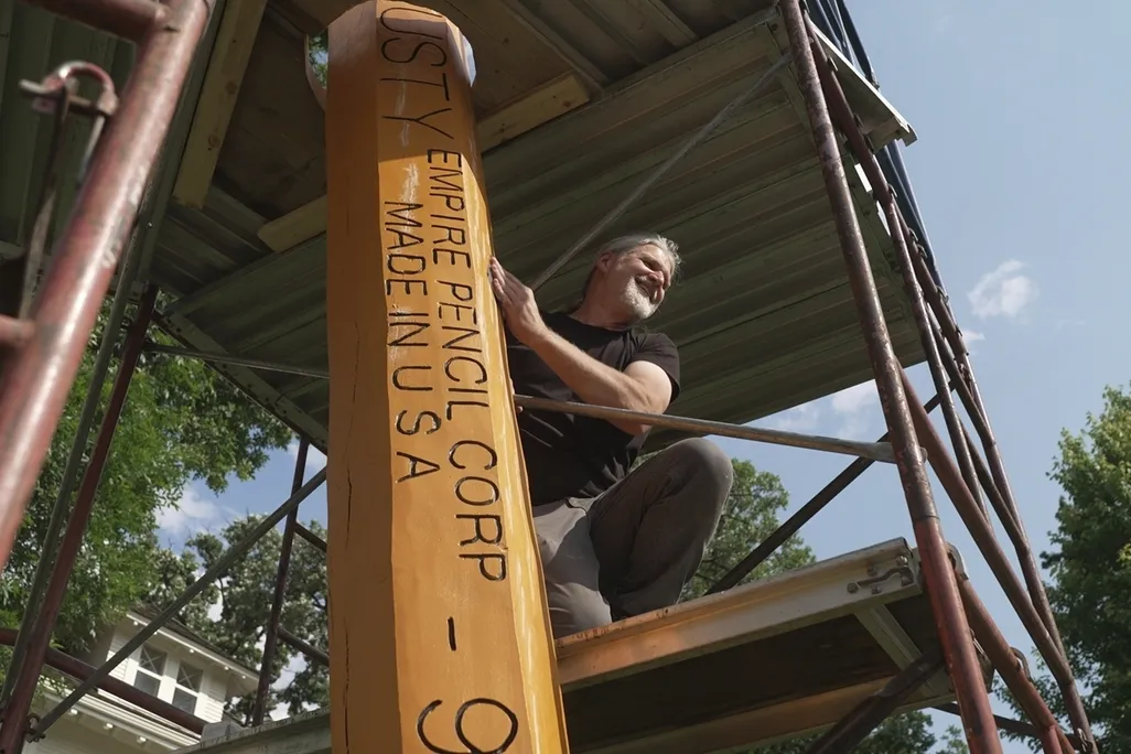 Man posing next to giant yellow pencil under scaffolding