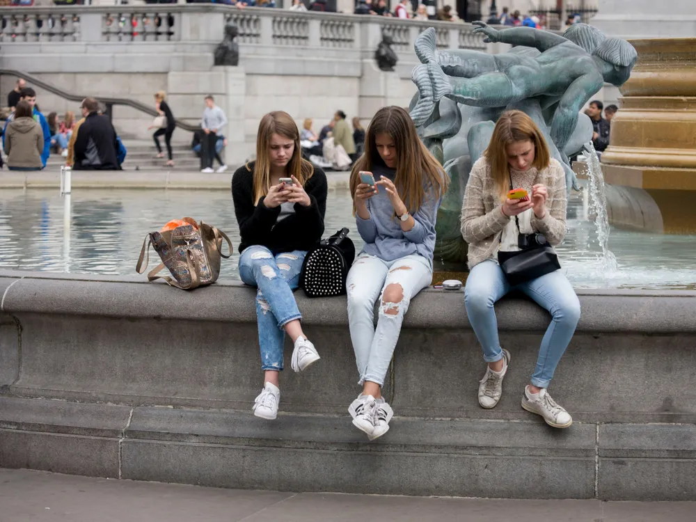 Three teenagers sit on the side of a large fountain and look at their smartphones