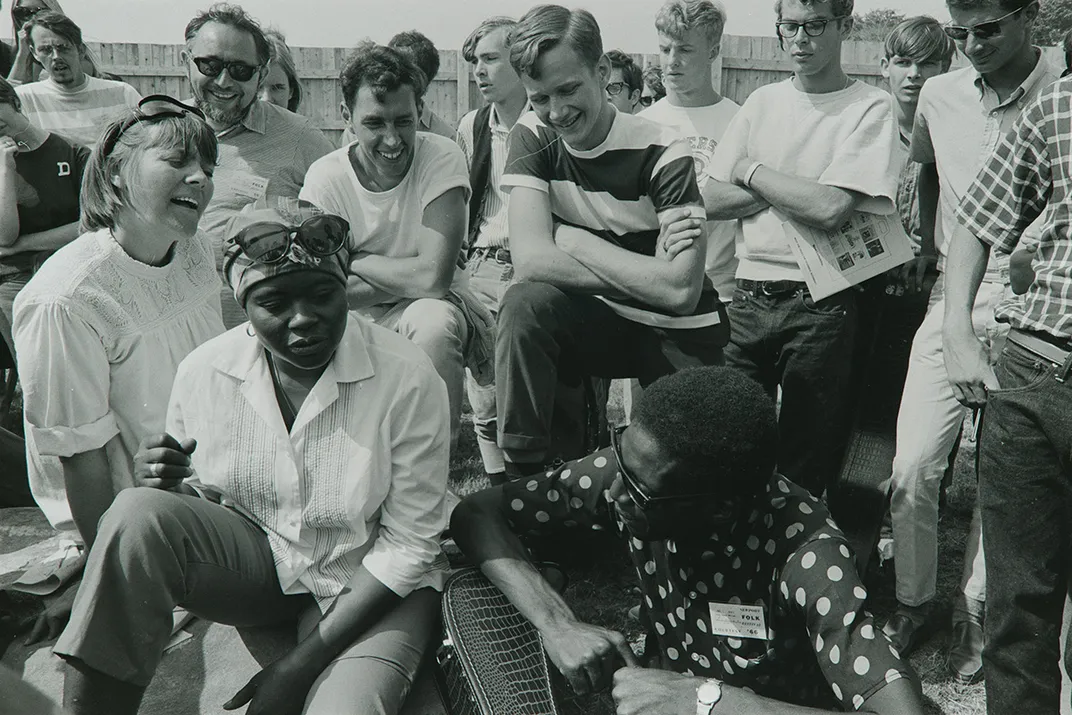 Black-and-white photo of a group of young people gathered outside, singing and smiling.