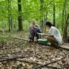 A young man and woman crouch on a wooden plank in a forest, with irrigation pipes across the ground. The man is pointing to a clear pipette in his hand.