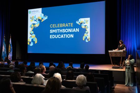 A woman stands at a podium in a theater with "Celebrate Smithsonian Education" projected on the screen