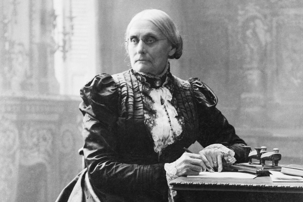 A black and white photograph of a woman with a tight bun, wearing a ruffled black dress with a white neckpiece, sitting a desk and in the middle of writing a letter, not smiling