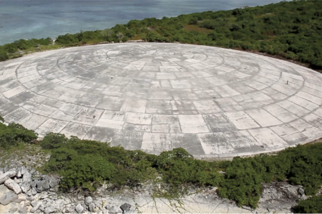 a large, circular concrete container on the coast, surrounded by trees and beach