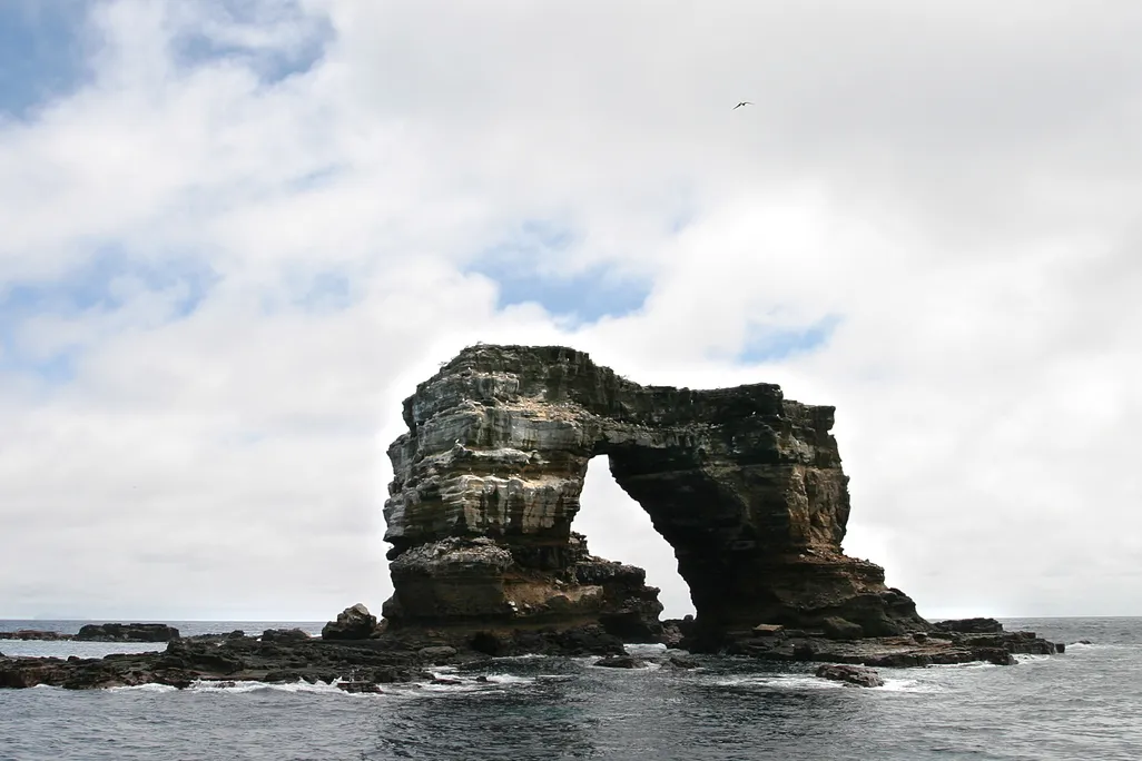 A photograph of Darwin's Arch in the Galàpagos Islands. The rock formation is two pillars attatched by a natural rock bridge. It stands itself in the ocean with a cloudy sky in the background.