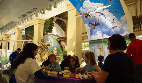 A group of children and two adults sit at a table creating origami birds among exhibits.