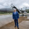 A 65-year-old man dressed in a blue suit poses in front of a military jet on a rain-soaked ramp.
