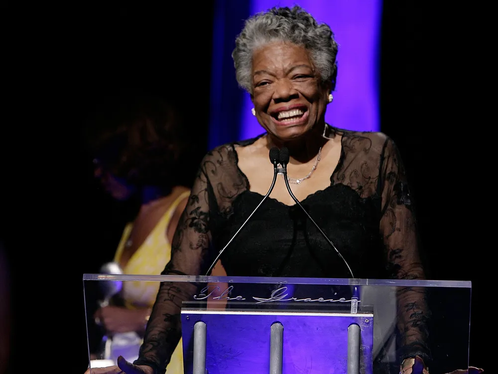 Angelou in front of a podium on a stage in a black formal dress, in front of a purple backdrop, smiling