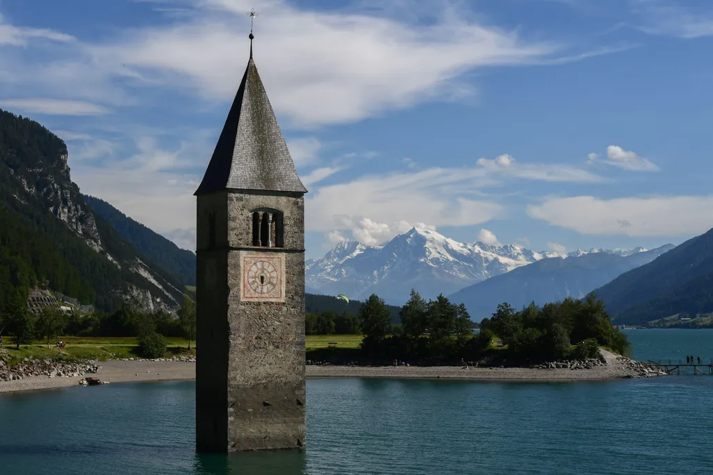 A picturesque view of a church steeple, old brick with a pitched roof, halfway submerged in water with a view of distant mountains and blue sky, wispy white clouds behind