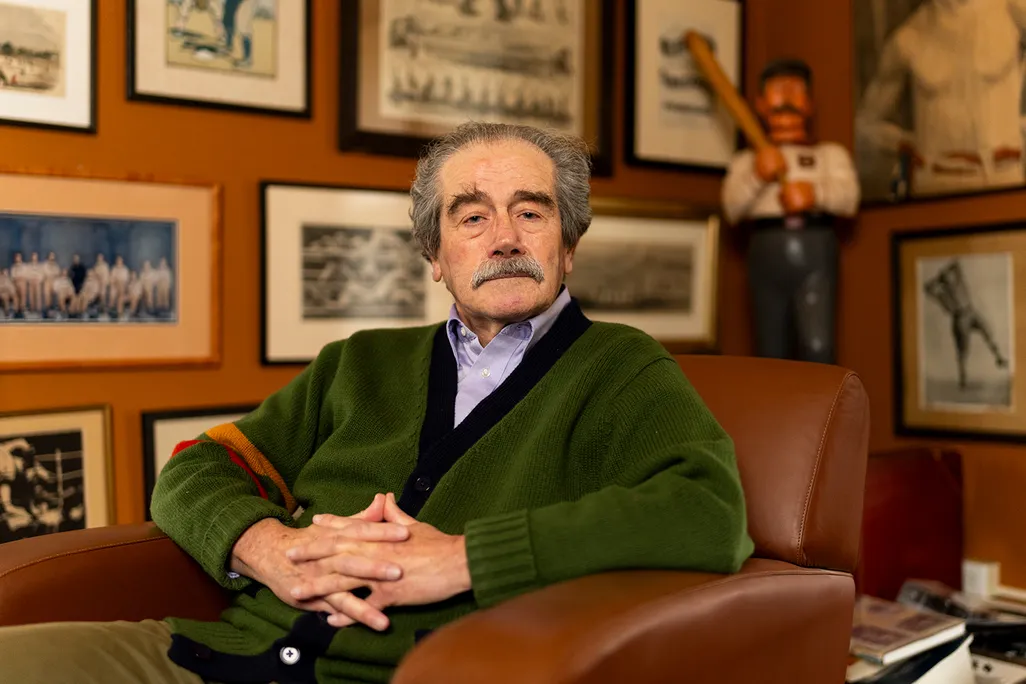 A man in a green sweater sits in front of a wall of hanging photographs