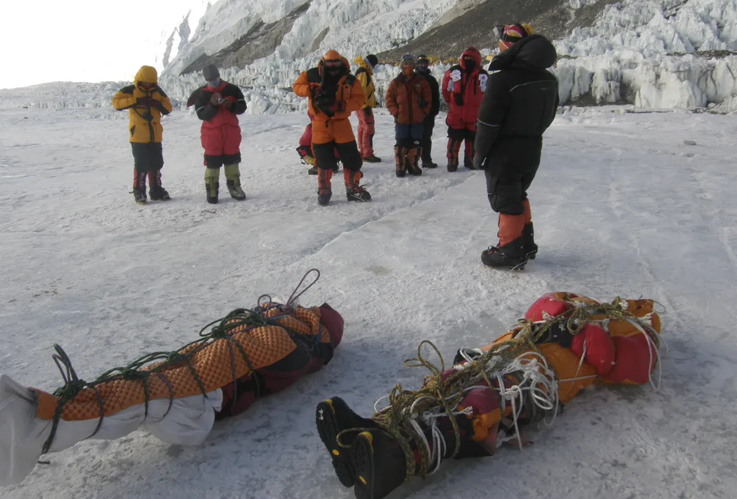 people in winter gear stand on a snowy mountain landscape with two bodies wrapped in rope at the front