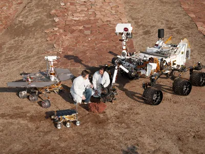 Scientists crouch with mock-ups of three generations of Mars rovers. Curiosity is the big one. Opportunity and Spirit were based on the medium-sized one on the left. The small one is front was the Sojourner rover.