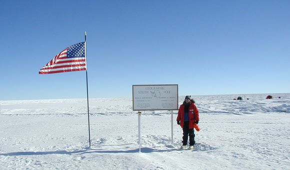 Geolgist Cari Corrigan poses for a picture in the snow next to an American flag and a sign that reads "Geographic South Pole."