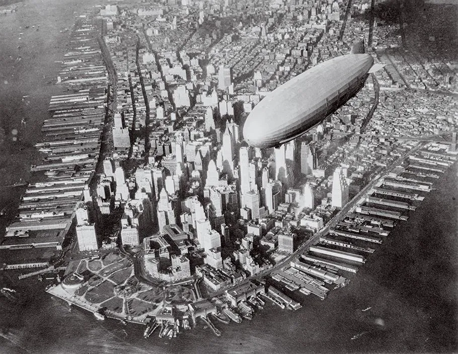 A black-and-white photo shows downtown Manhattan skyscrapers from above with a white airship edging into the image.