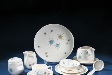 Photographed against a dark blue background is a studio image of a set of cream-colored ceramic dishware bearing a pattern of star-shapes in blue, yellow, and green. There are several pieces, including a platter, a gravy boat with a ladle, a creamer and s