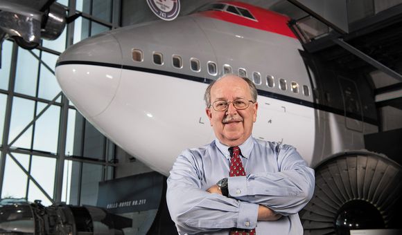 A white man in his early 70s stands inside a museum gallery. He is wearing glasses and smiling. Behind him, is the nose and forward fuselage of a Boeing 747 airplane. The 747 is painted in a red, gray, and white paint scheme.