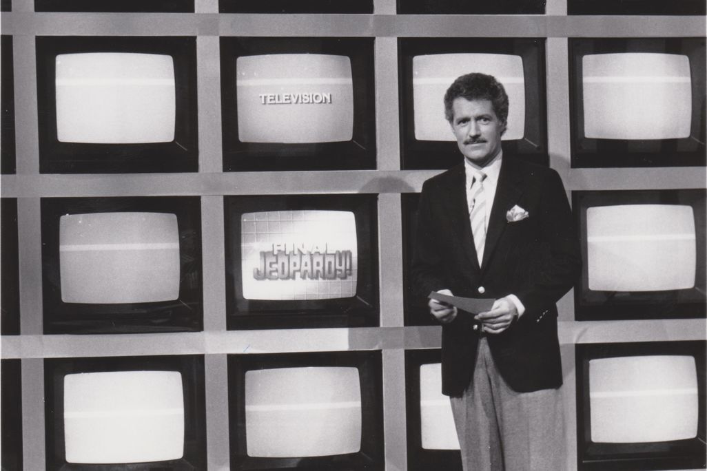 A black and white image of a young Trebek, a white man in a suit and tie with dark hair and a mustache, standing in front of a grid of TV screens