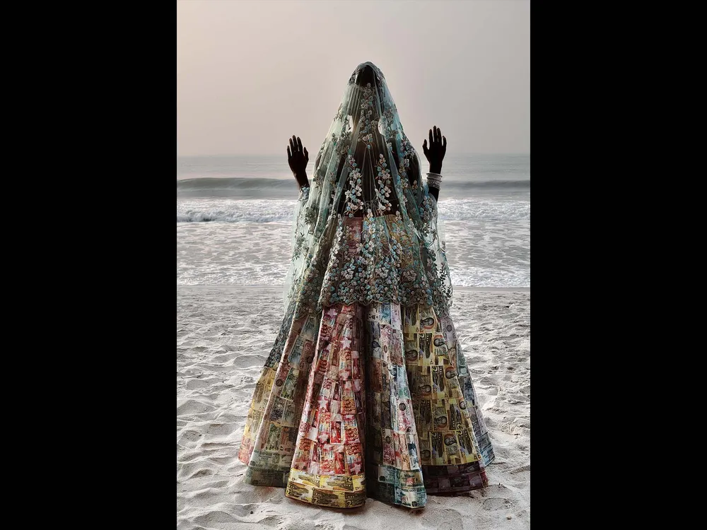a person stands  in a decorated dress and vail on a beach with the sea behind them