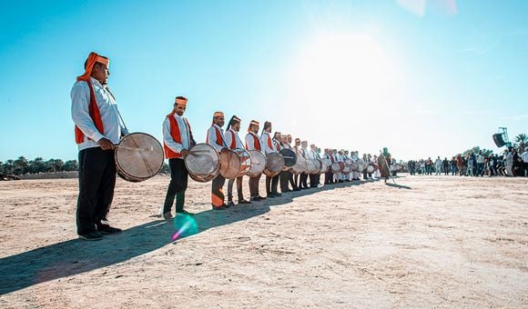 A line of drummers wearing red vests and white shirts stand in the dessert on a clear, blue-skied day.