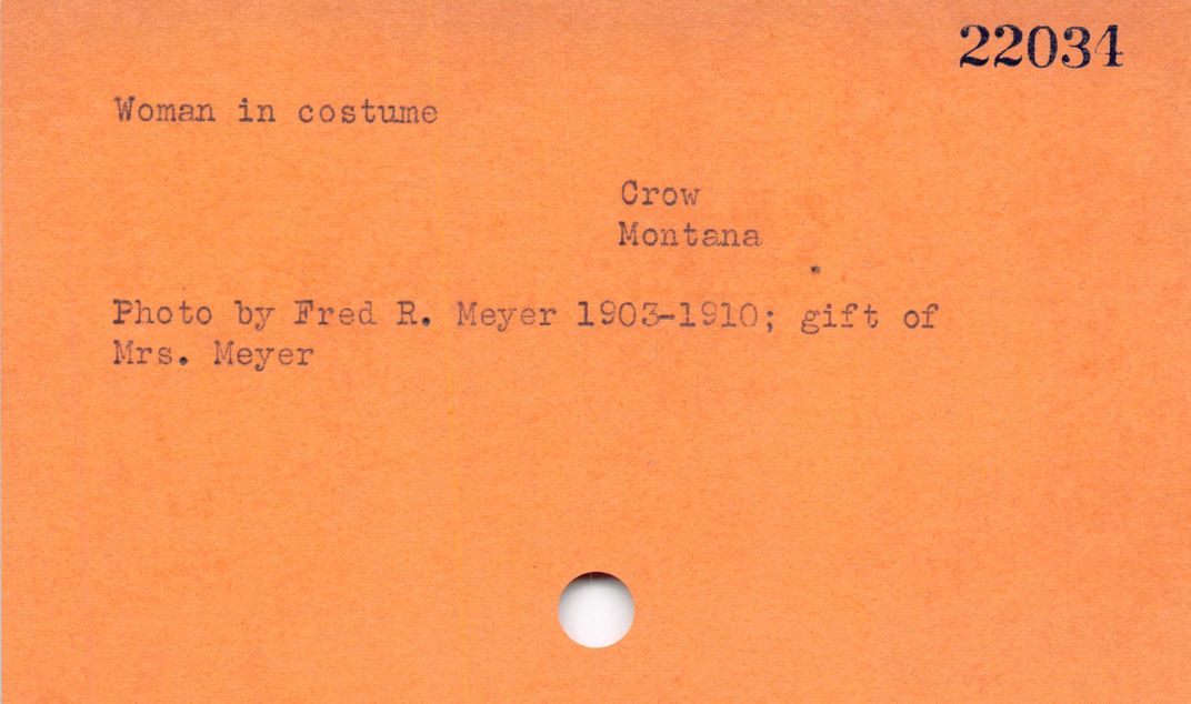 Orange card with a single hole punch at the bottom with black text " 22034 Woman in costume Crow Montana Photo by Fred R. Meyer 1903-1910; gift of Mrs. Meyer"