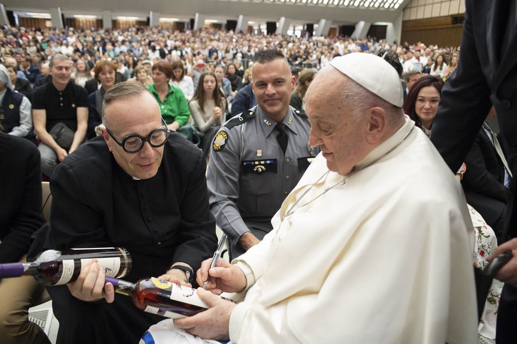 A man wearing glasses holding a bottle next to a man wearing a white robe and a little white hat signing a bottle