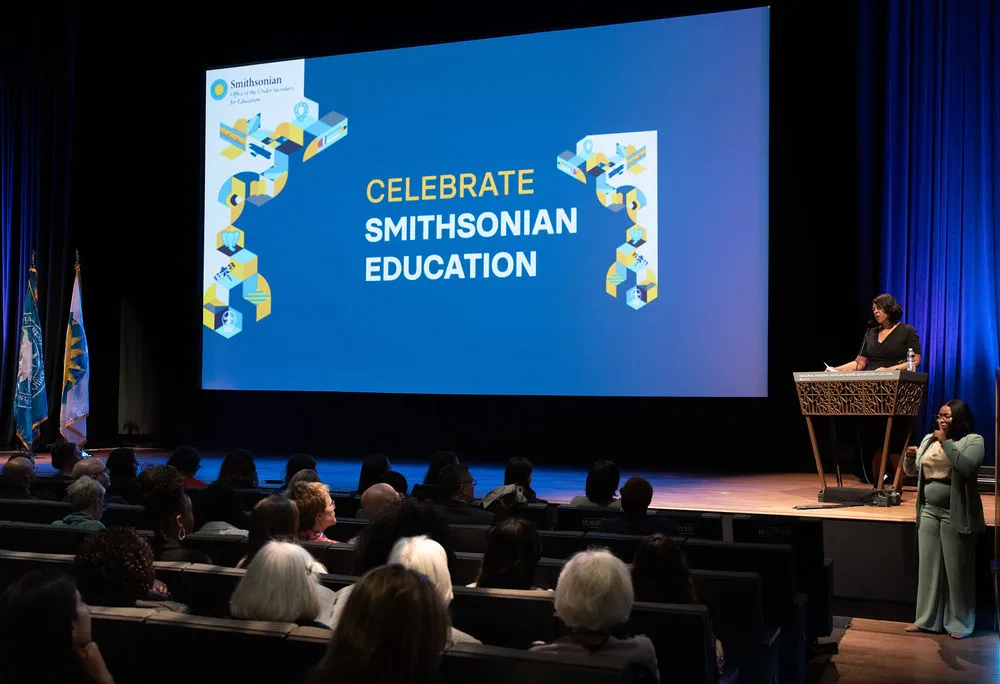 A woman stands at a podium in a theater with "Celebrate Smithsonian Education" projected on the screen