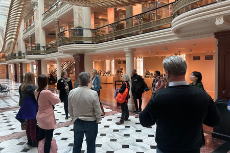 People stand and listen to a speaker in a brightly lit museum room underneath a large gold art installation.
