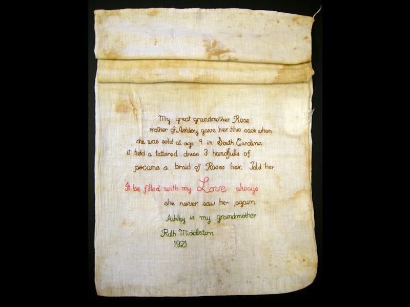 A yellowed, stained sack made from pale white cotton, with its top two thirds roll and its lower segment extended to reveal an embroidered message, stitched in elegant looping script