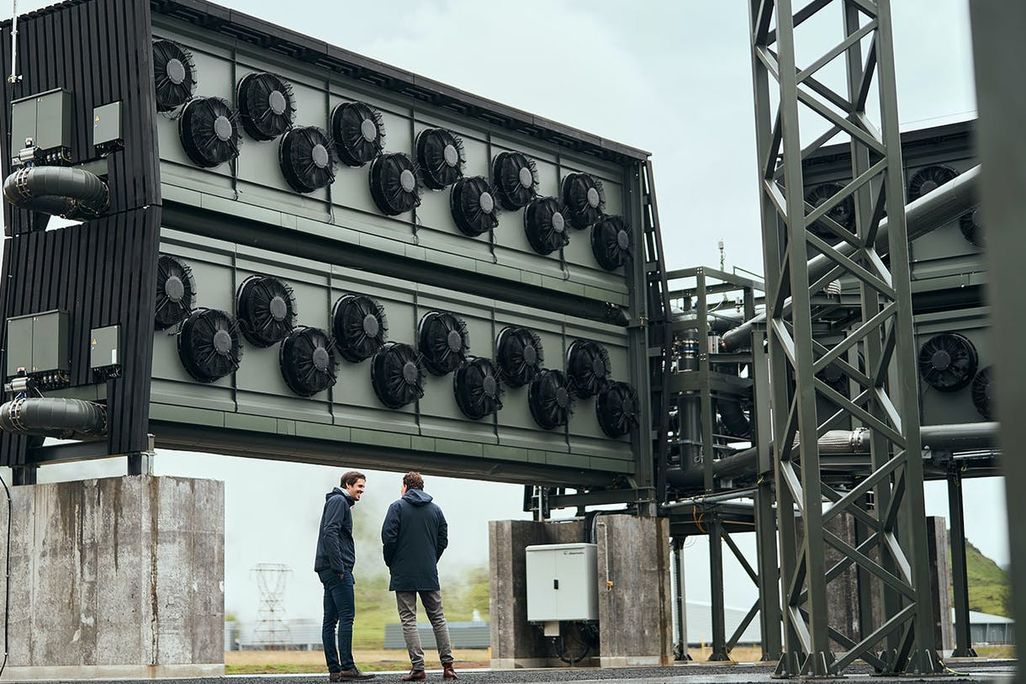 Two men stand in front of tall wall of fans at the Orca carbon capture facility in Iceland
