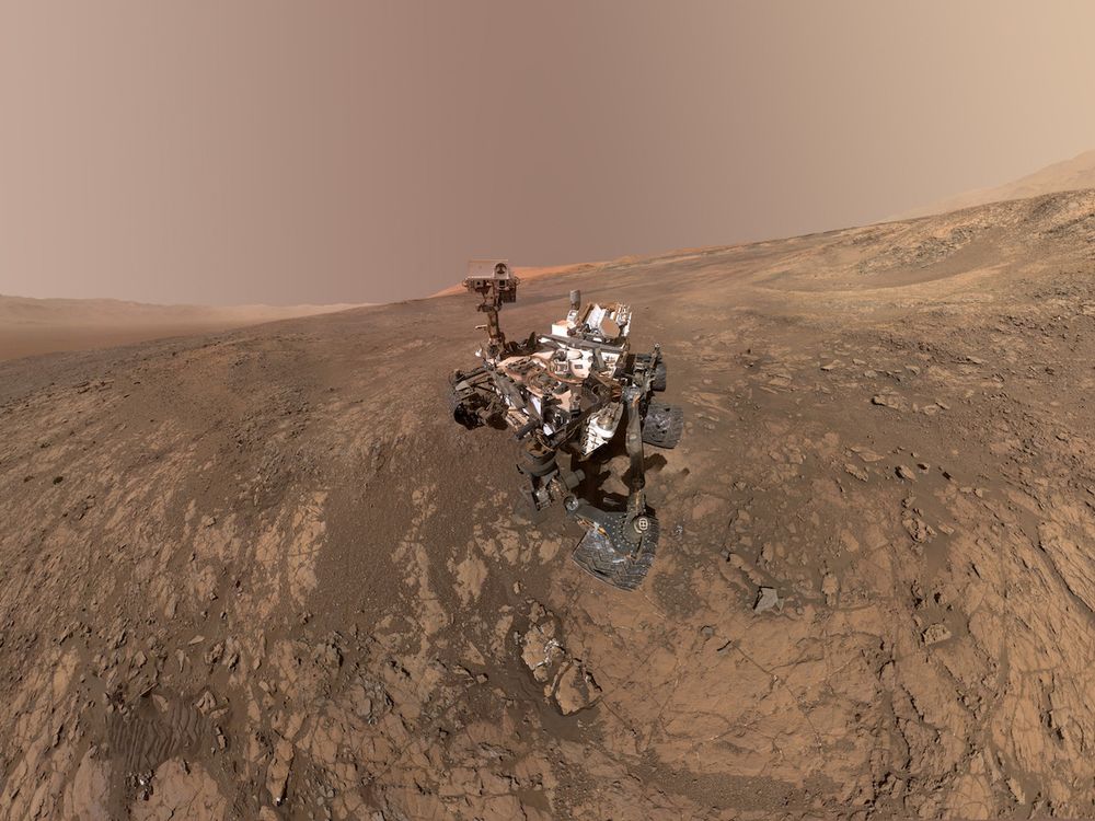 An image of the Mars Curiosity rover on planet Mars with Mt. Sharp in the background. On the right, towards the horizon is the Gale Crater's rim.
