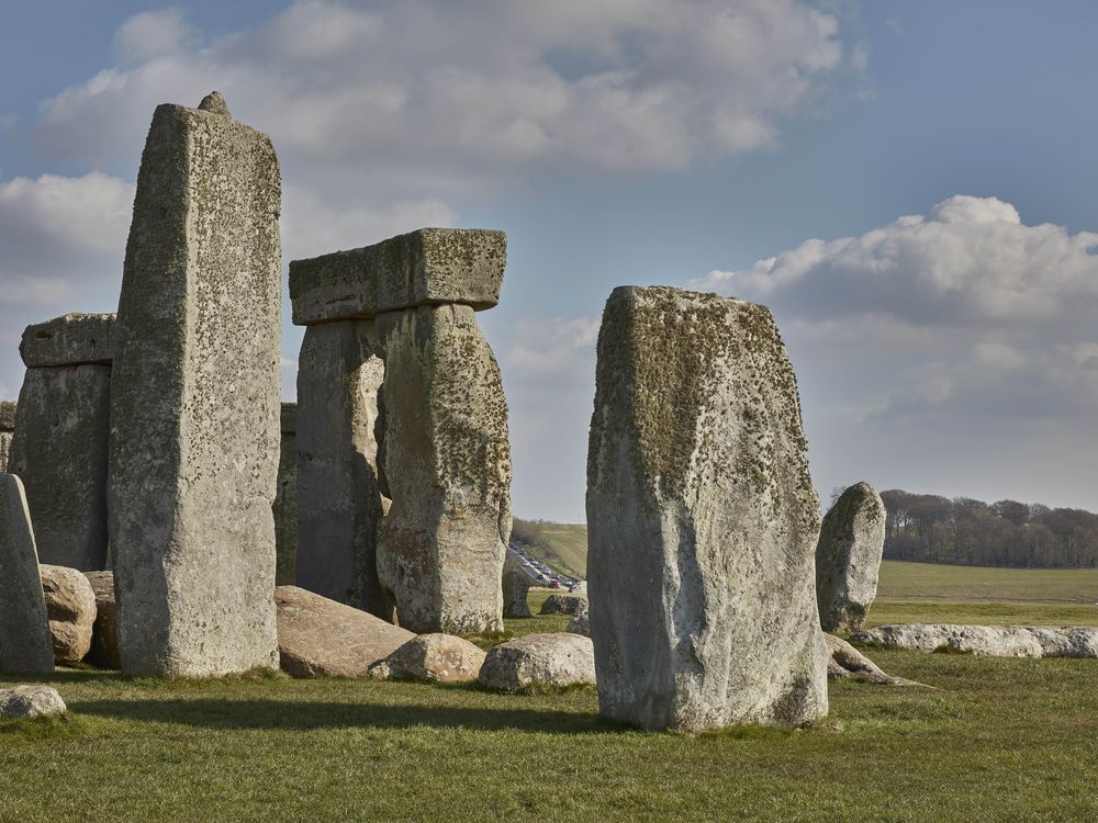 A view of Stonehenge's iconic square stones, under a blue sky with puffy white clouds; in the distance between two of the large stones, a trafficked road full of cars snakes over a hill