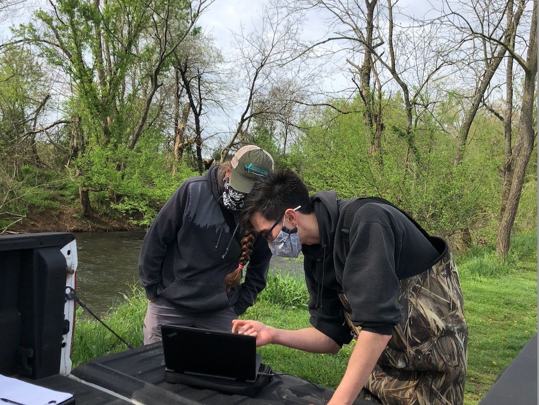 Researchers wearing masks examine something on a computer screen while standing near a river.