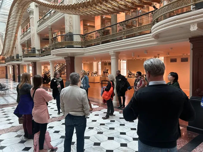 People stand and listen to a speaker in a brightly lit museum room underneath a large gold art installation.