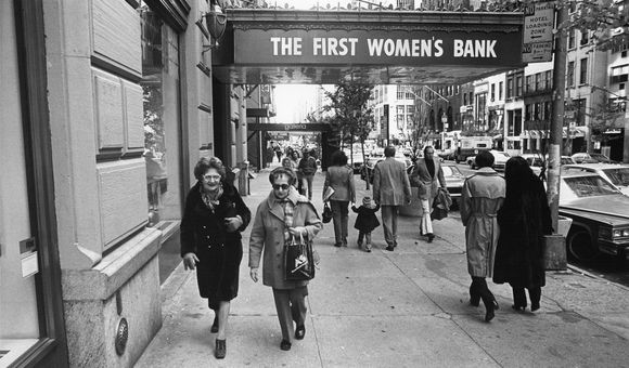 Candid black and white photo of people walking down the street. Two women face the camera in the foreground under an awning with the words “The First Women’s Bank” on it.