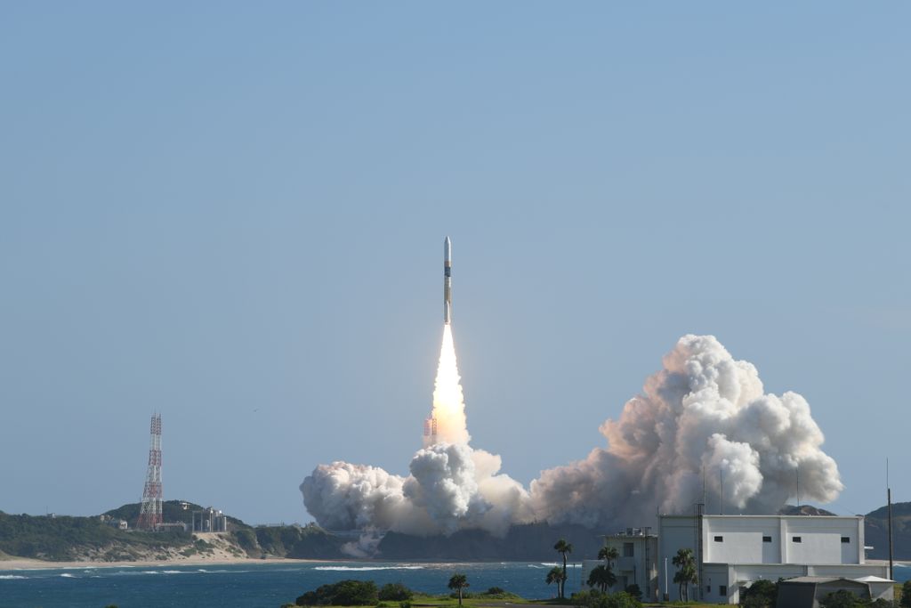 In the distance, a rocket launches into a clear sky over clouds of smoke